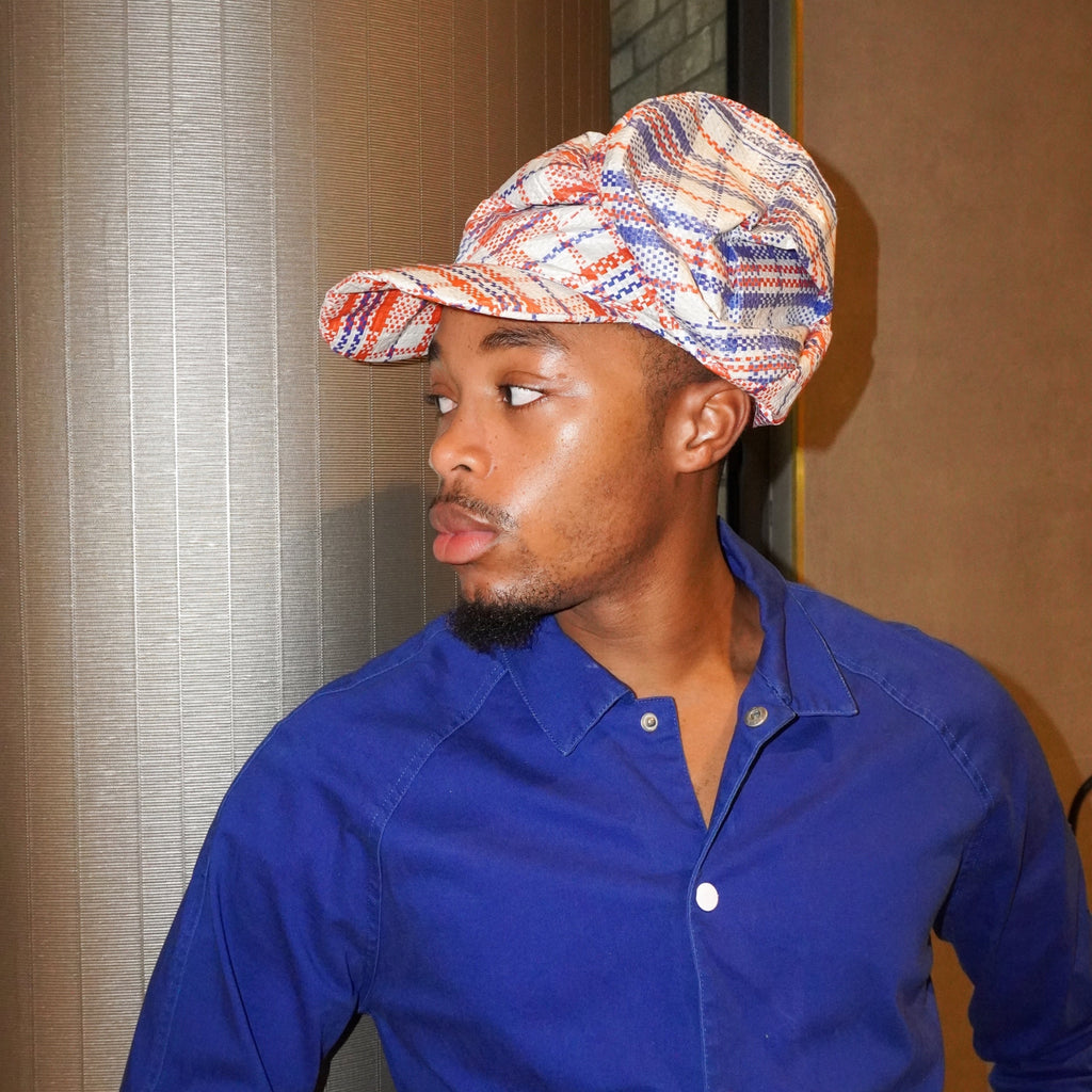 A man wearing a blue button-up shirt and a patterned cap with red, blue, and white plaid details, standing indoors against a textured beige wall.