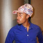 A man wearing a blue button-up shirt and a patterned cap with red, blue, and white plaid details, standing indoors against a textured beige wall.