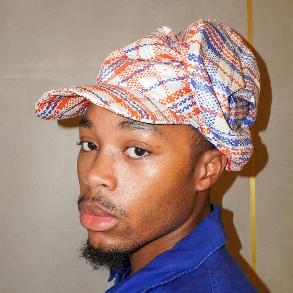 A man wearing a blue collared shirt and a patterned cap with red, blue, and white tones, photographed against a plain beige wall.