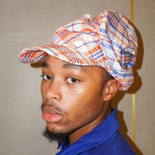 A man wearing a blue collared shirt and a patterned cap with red, blue, and white tones, photographed against a plain beige wall.